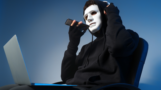 Cilvēks melnā apģērbā ar kapuci galvā. Uz sejas balta maska. Tur rokās tālruni, kurš pielikts pie mutes. Klēpī portatīvais dators.
