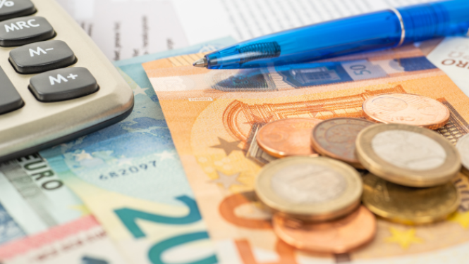 Uz galda novietots kalkulators, zila pildspalva, papīra nauda un monētas