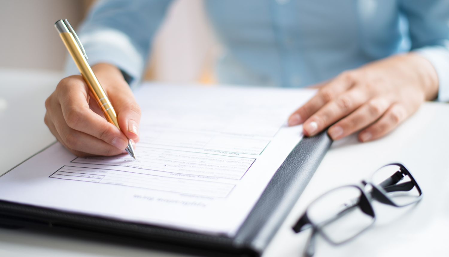 brilles, dokumentu mape un rakstoša cilvēka roka
