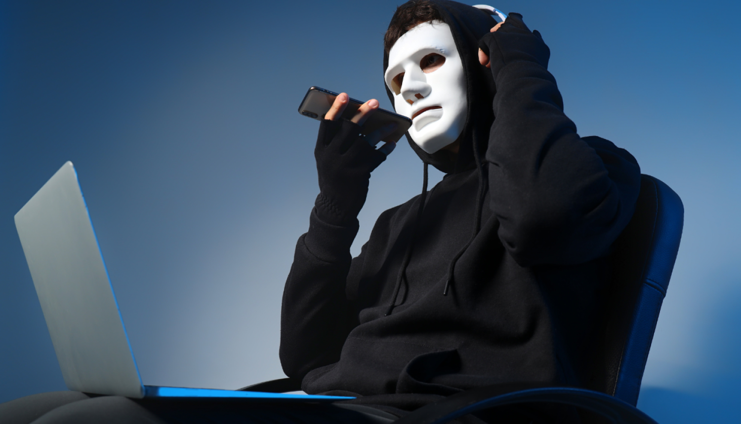Cilvēks melnā apģērbā ar kapuci galvā. Uz sejas balta maska. Tur rokās tālruni, kurš pielikts pie mutes. Klēpī portatīvais dators.